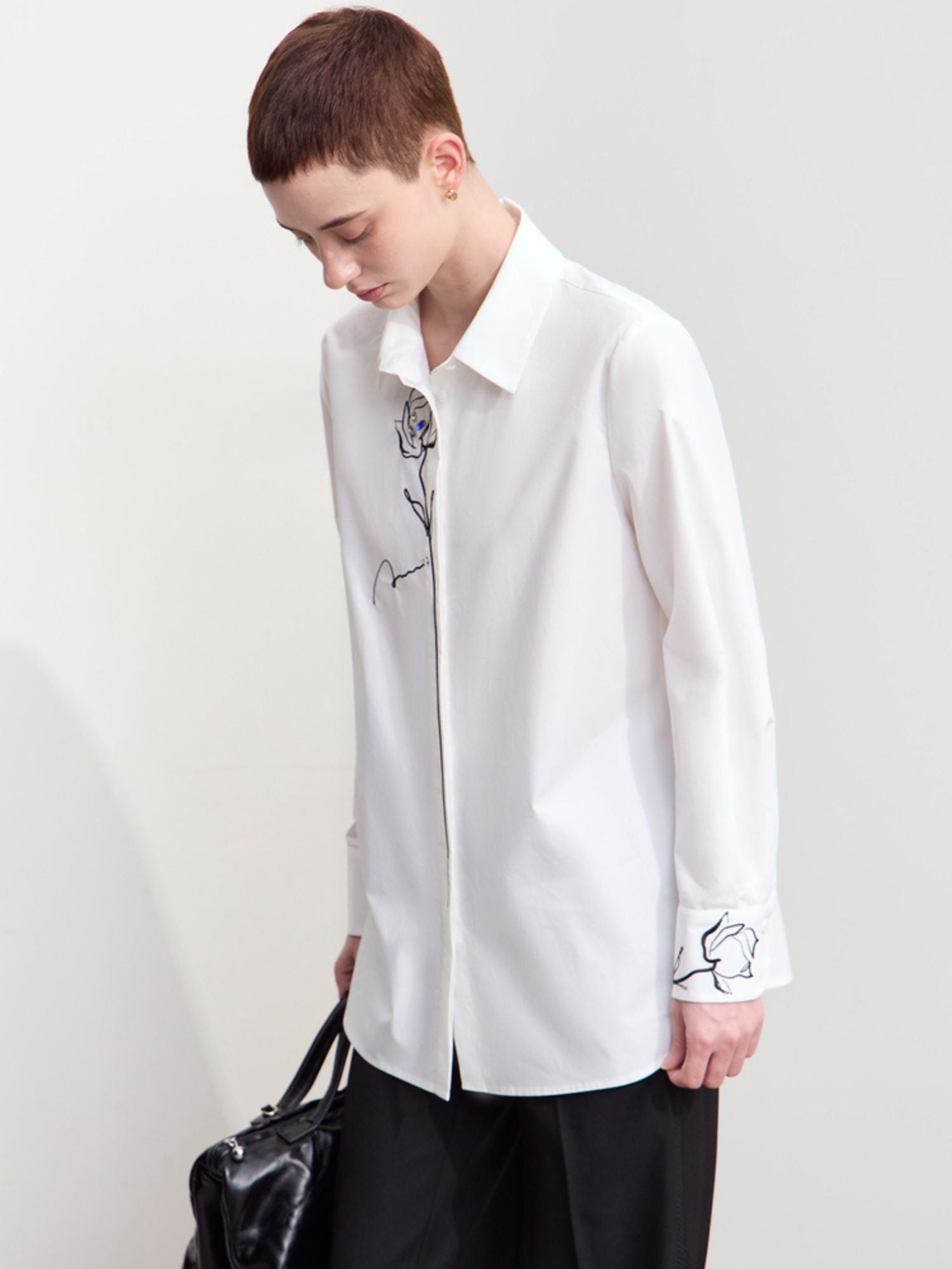Side view of model in white embroidered shirt and black pants, holding a black bag