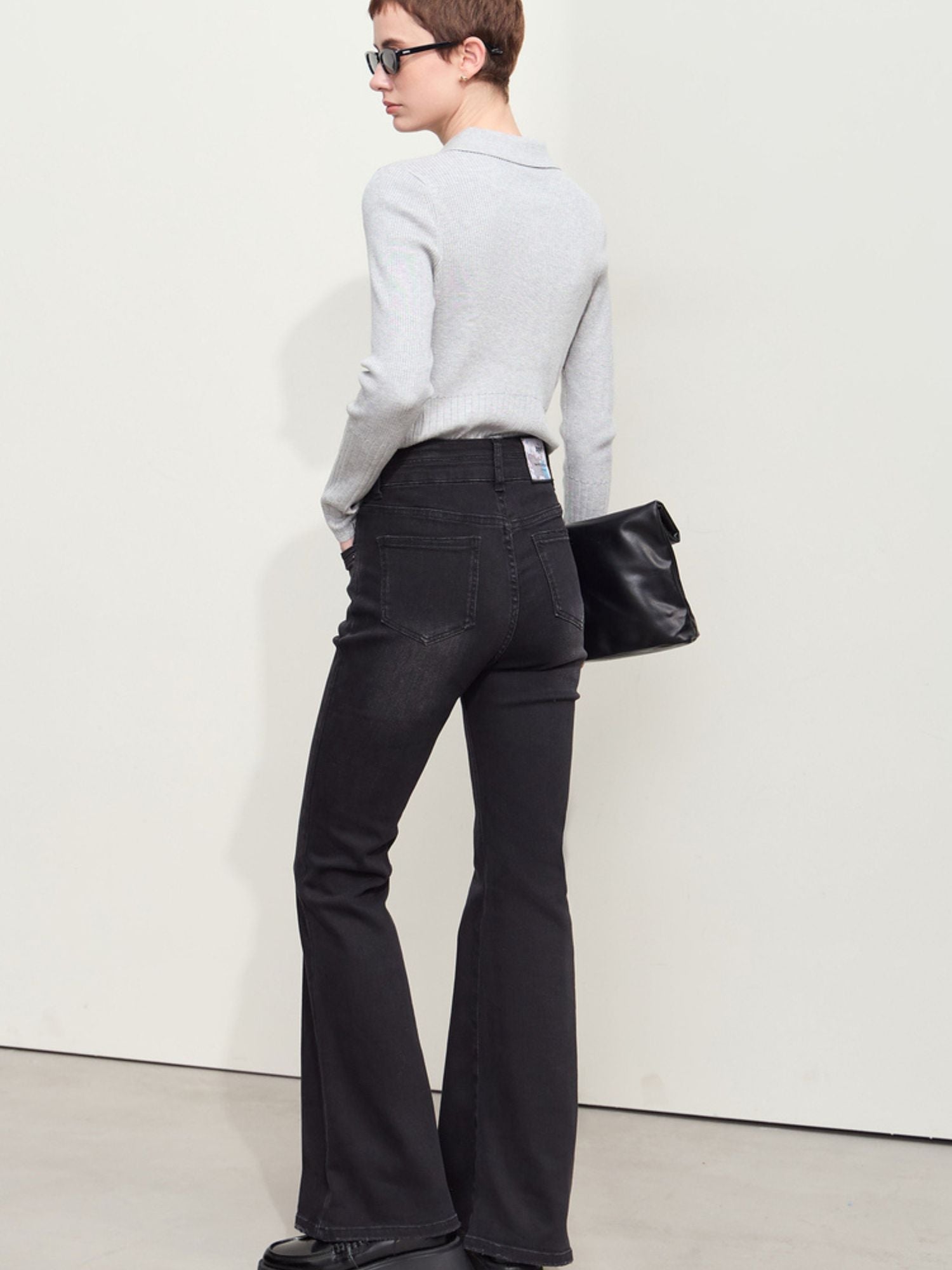 Whole body and a front view of a person wearing high-waisted, flared black jeans paired with black bag and shades