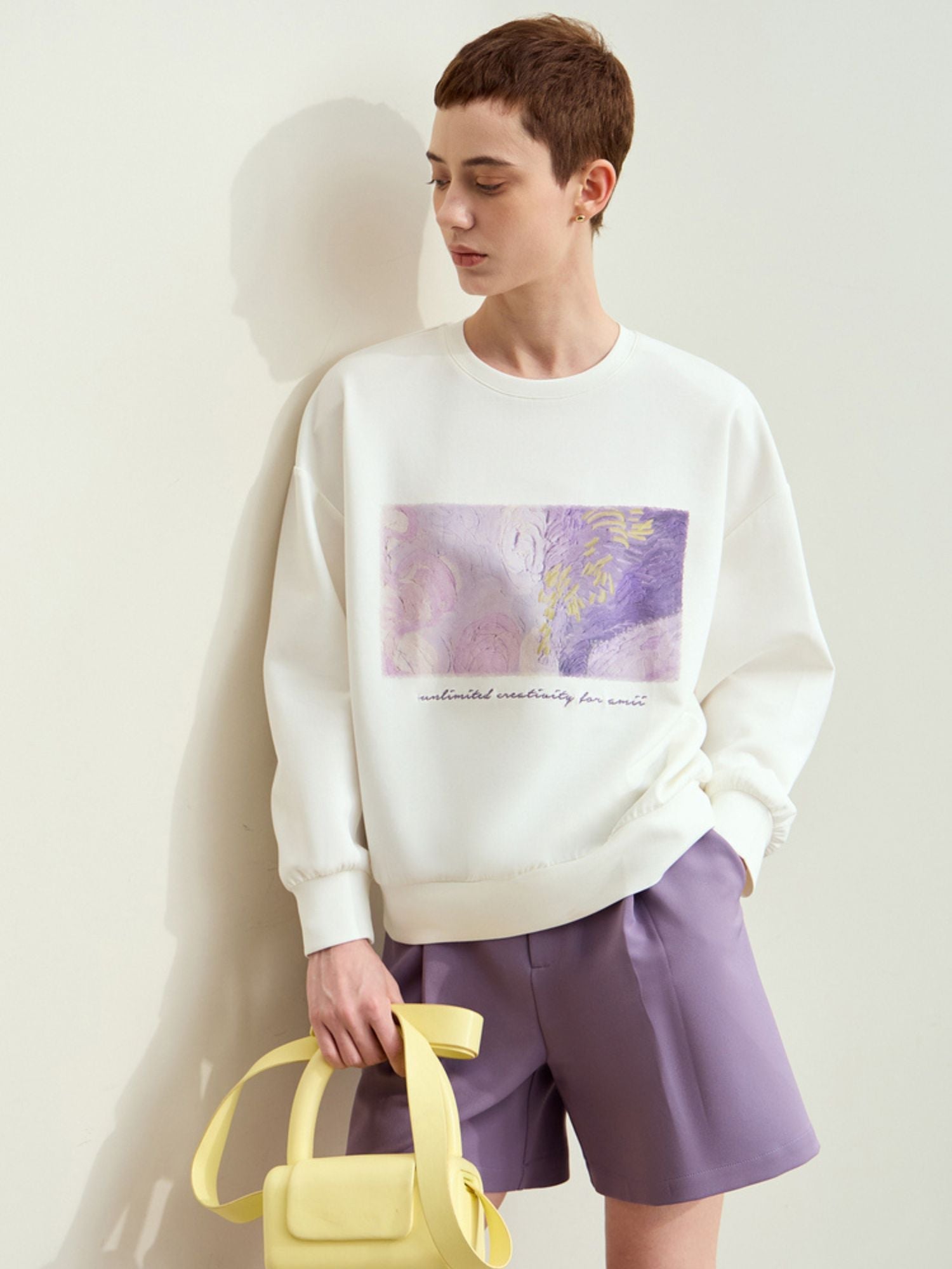 Model wearing white sweatshirt with abstract purple graphic paired with lavender shorts, accessorized with yellow bag