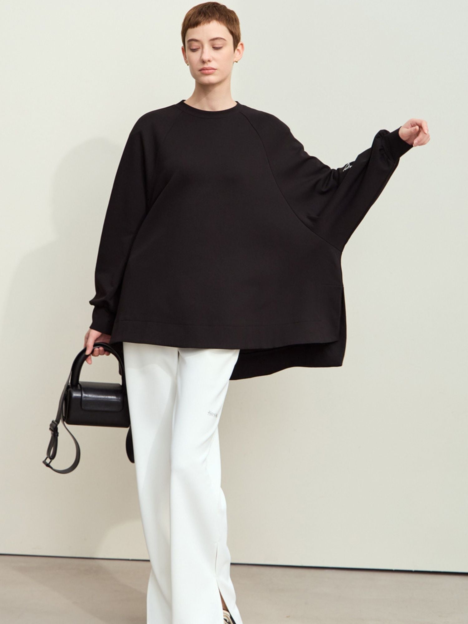 Model lifting one arm while wearing a black oversized sweatshirt and white pants and a black handbag