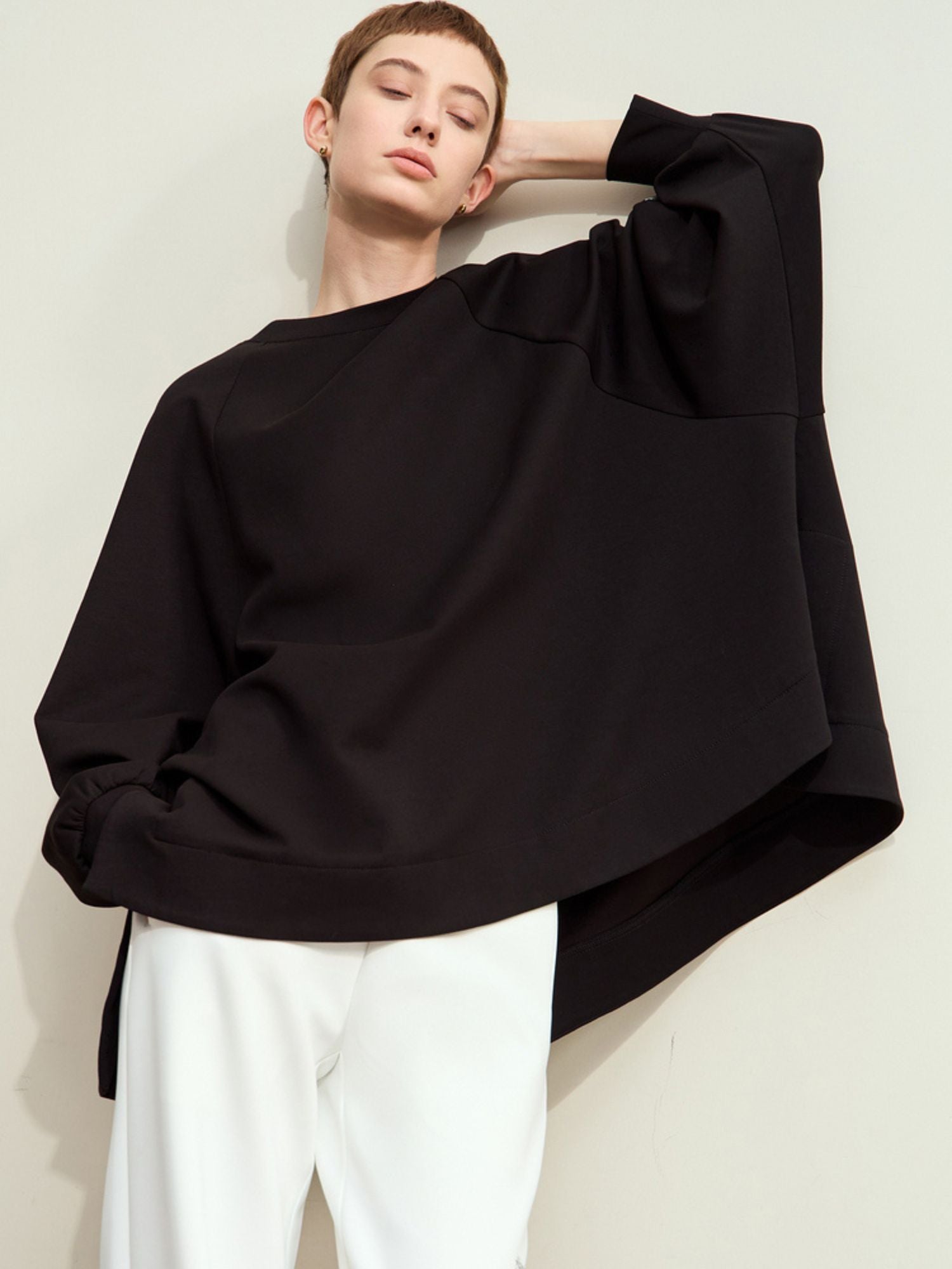 Model lifting one arm while wearing a black oversized sweatshirt and white pants, cropped at hip
