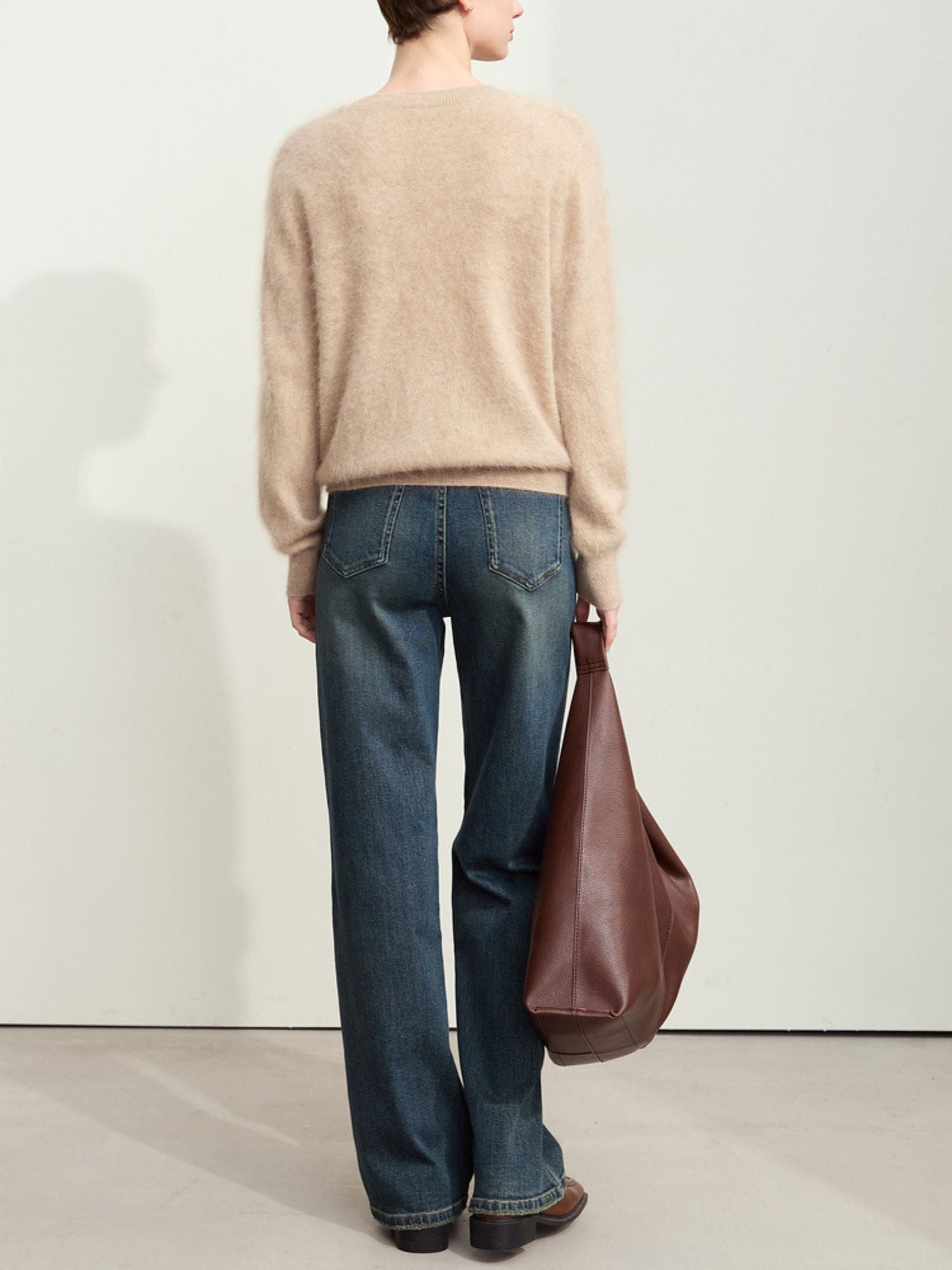 Back view of a person wearing straight-leg jeans, a beige sweater, and holding a brown bag