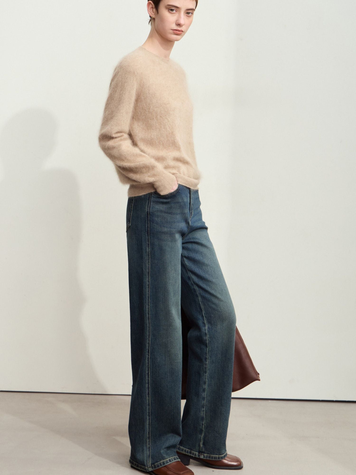 Side view of a person in straight-leg jeans and a beige sweater, with brown shoes and a large brown bag