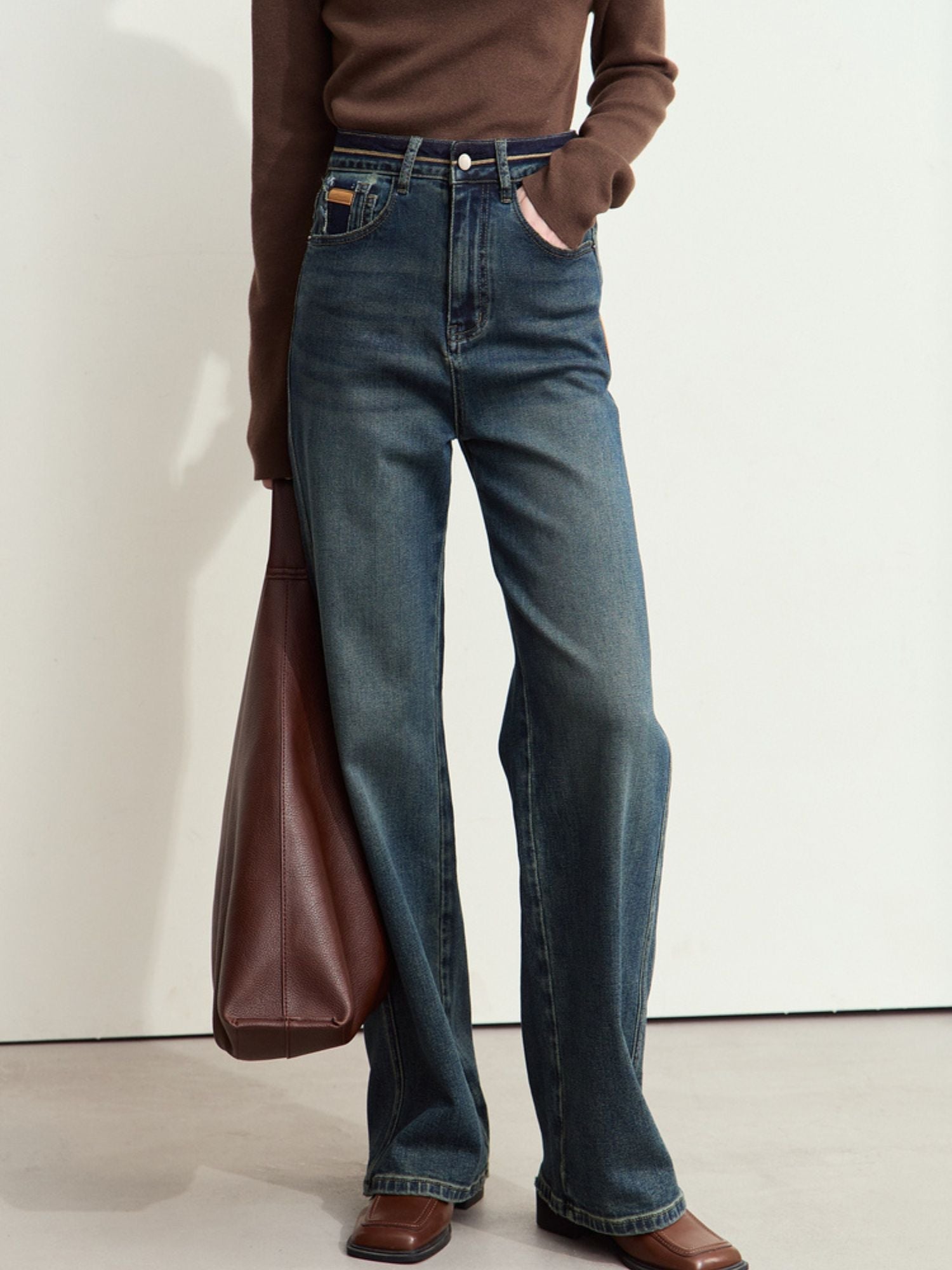 Three-quarter front view of a person wearing blue jeans, brown shoes, and a brown bag resting against the leg